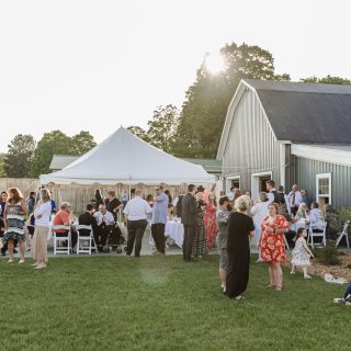 A large wedding crowd on the patio and in the courtyard of The Blue Ribbon Barn enjoying a wedding reception on a sunny day. Some are sitting under the tent to enjoy shade.