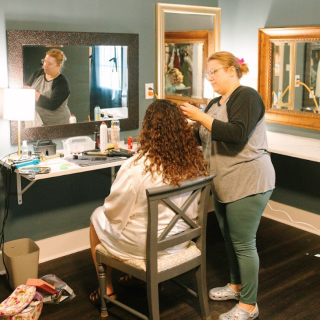 A bride getting her hair done by a wedding hair stylist in The Blue Ribbon Barn bridal lounge. She is sitting in front of multiple mirrors with good lighting.